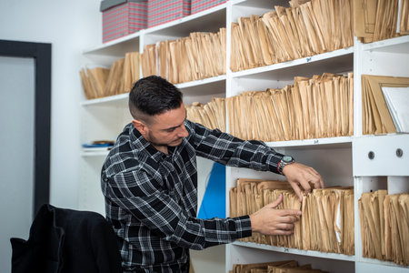 Office worker searching through files in an archive room, organizing paperwork and retrieving important documents from shelvesの写真素材