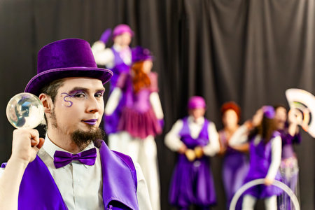 Magician wearing purple costume and top hat holding crystal ball performing with circus troupe in backgroundの写真素材