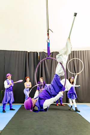 Male acrobat wearing purple costume hanging upside down performing on aerial hoop with other circus performers in background on stageの写真素材