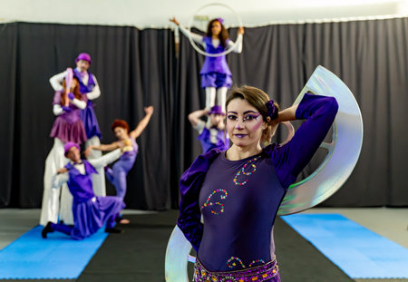 Confident circus artist posing with large semicircular props, while other performers practice acrobatics and balancing acts in the backgroundの写真素材