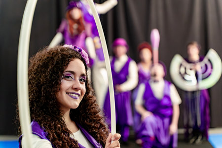 Young woman with curly hair and purple makeup smiling and holding a hula hoop, with other circus performers blurred in the backgroundの写真素材
