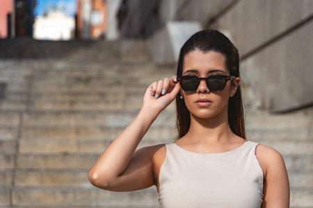 Stylish young woman confidently posing on outdoor stairs, adjusting her sunglasses with an effortless blend of modern elegance and casual flairの写真素材
