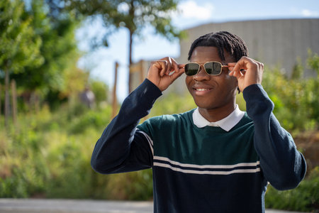 Smiling african american student putting on sunglasses in a university campus park, enjoying a sunny dayの写真素材