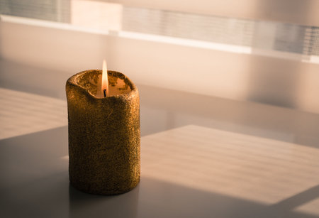 Burning candle on a wooden table next to a window through which a faint evening light entersの写真素材