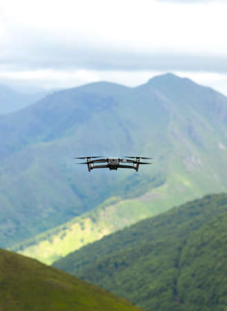 Drone flies away over a mountainous landscape. The drone can be seen from behind flying away. In the background you can see a mountain and below it a forest and a green meadow.の写真素材