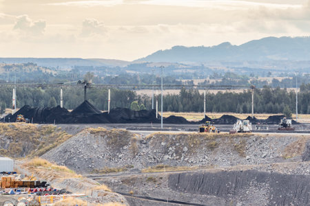Coal mining scene showing large mining trucks, excavators, and black coal deposits under a wide...