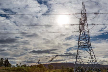 Sun shining through a cloudy sky, illuminating a lattice power tower with high-voltage power lines....
