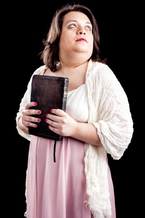 hispanic looking woman in a light dress against a black background holding a bible close to her body as she looks up in thought. の写真素材