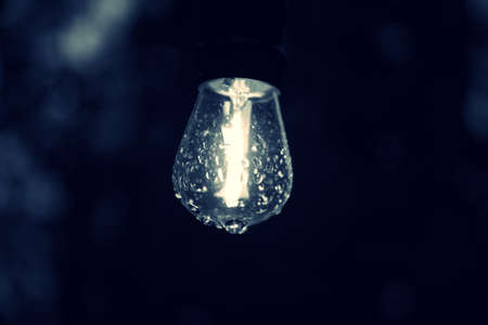 Close up on outdoor lit lightbulb with droplets of rain on the glass.