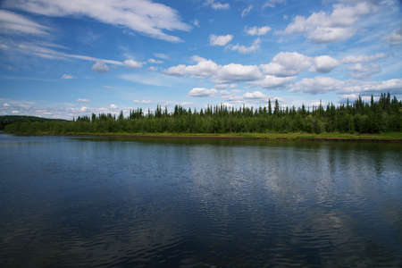 Forest on Riverbank with blue sky and clouds reflected in the water.の写真素材