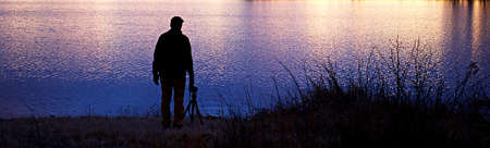 Silhouette of person standing by lake, with golden sunlight reflected in blues, purple, yellow, and pink on the water.の写真素材