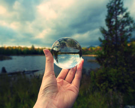 Hand holding crystal ball against horizon reflected landscape of river, pine trees, and overcast...