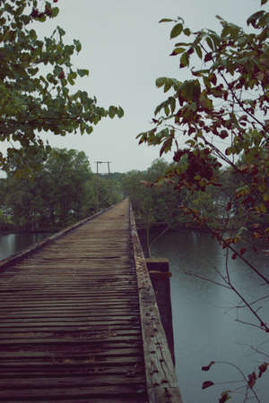 Old wooden bridge once part of railroad track, crossing a river with forest on both sides.の写真素材