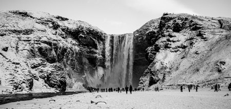 Waterfall SkÃ³gafoss on a winter day with snow-covered fields and people getting an up-close view of the waterfall.の写真素材