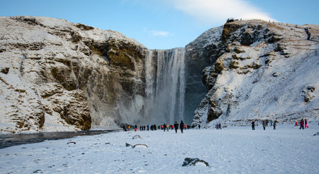 Waterfall SkÃ³gafoss on a winter day with snow-covered fields and people getting an up-close view of the waterfall.の写真素材