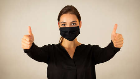 Positive woman wearing black face mask showing her thumbs up. Happy young Caucasian girl expressing encouragement and support with her fingers during medical crisis of coronavirus.の写真素材
