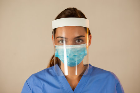 Caucasian female with attached face shield with light background. Medical worker with protective equipment looking into camera. Female in blue shirt with face mask on.の写真素材