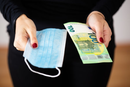 Concept of coronavirus crisis costing money and creating economical problems. Woman holding blue surgical face mask in one hand and green one hundred euro bank note in other. Expensive healthcare.の写真素材
