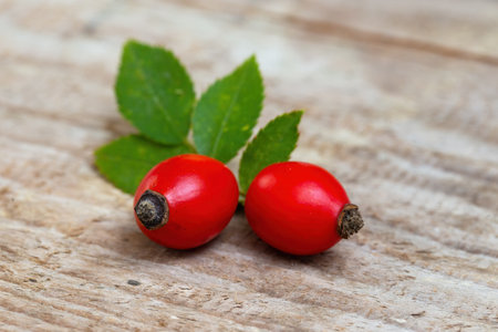 Red rosehip berries lying on wooden table from close-up. Two ripe brier seeds with green leaf on brown background. Detailed view on raw food rich in vitamin c.の写真素材