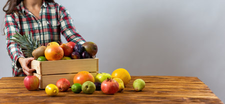 Young lady putting wooden box with fruit on the table. Woman in flannel shirt touching wood crate full of fresh sweet food on the brown desk with others plants around. Person with tasty meal.の写真素材