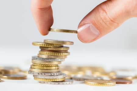 Fingers putting one euro on pile of coins from close up. Concept of saving money for future. Adding...