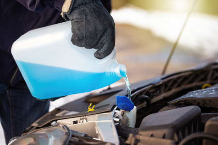 Man filling antifreeze coolant for cleaning front window with snow in background. Concept of winter maintenance of car. Person replacing blue liquid in engine.の写真素材