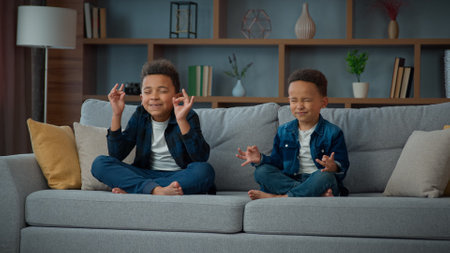 Two funny little African American ethnic children boys brothers siblings kids meditating together sitting on couch in lotus position. Small schoolboys meditate. Spiritual development yoga meditationの写真素材