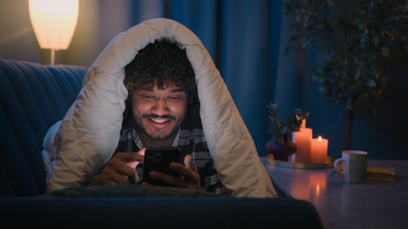 Smiling guy Indian man Arabian male at night evening home on couch under blanket cover with duvet...