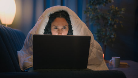 Hispanic freelancer girl Latino woman Indian Arabian female at night evening home at couch sofa under blanket cover duvet working with laptop watching movie at computer browsing social media addictの写真素材