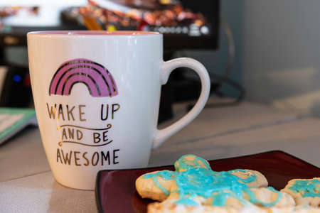 Wake Up and Be Awesome coffee cup in focus with glazed sugar cookies on a red plate in foreground out of focus and  computer monitor in background out of focus.の写真素材
