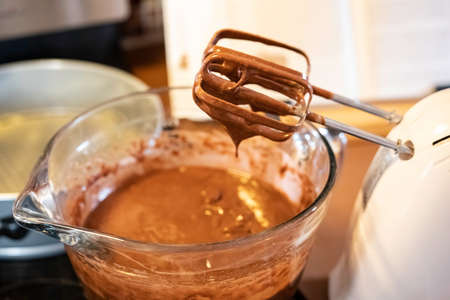 Chocolate cake batter dripping off of the hand mixer into a glass mixing bowl with baking pan and cookbook in background out-of-focus.の写真素材