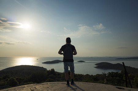 Middle age caucasian man with bag on back standing outdoors on sunny spring day looking at Adriatic Sea in Croatia. Beautiful nature and landscape of Dalmatia coast. Nice sunset at the ocean.の写真素材