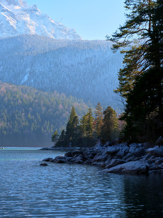 Tranquil lakeside view with mountains and evergreen trees at sunrise in a natural landscape settingの写真素材