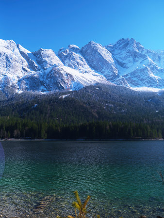 Snow-capped mountains reflecting in the clear lake under a bright blue sky during a sunny day in early winterの写真素材