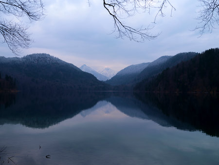 Serene lake reflecting majestic mountains under a cloudy sky in a tranquil winter landscapeの写真素材