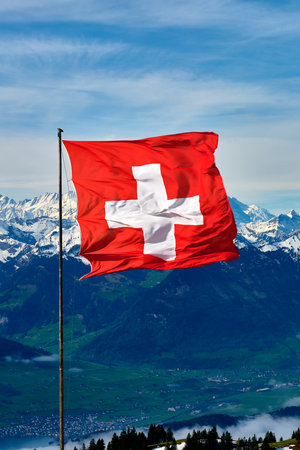 Swiss flag waving proudly against a backdrop of majestic mountains and a clear blue skyの写真素材