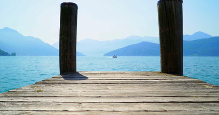 View of a wooden dock extending over a lake surrounded by mountains on a clear day with a boat in...