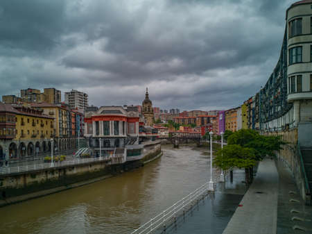 Cityscape of Bilbao, Spain, with the market hall and the spire of the church San Anton aside the river Nervion with colorful buildings and a dark cloudy skyの写真素材