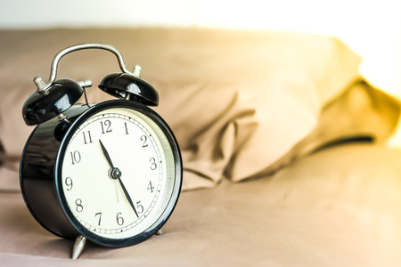 Black cover alarm clock is putting on a bed with pillow behind in brown cover with yellow sun light at lately morningの写真素材
