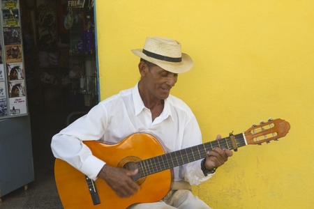A man is playing the acoustic guitar and sings in a street of Trinidad, Cuba. He wears a white shirt and a hat and is seated in front of a yellow wallのeditorial素材