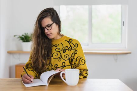 Woman with glasses writing with a pencil on a notebook. A white mug on top of a wooden workbench...
