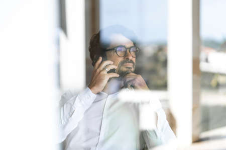 Man wearing glasses making a mobile phone call sitting down looking through the window. Successful...