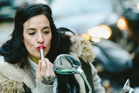 Beautiful brunette woman, middle age, painting her red lips in a mirror of a motorcycle.の写真素材