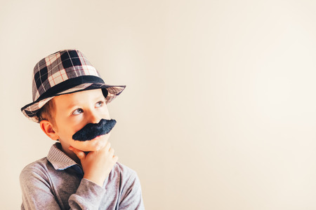 Funny portrait of pensive boy with fake mustache and hat, concept of childish humor, copy space.