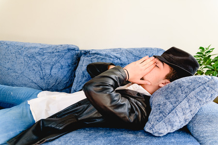 Young man tired of working makes a break for a nap on a sofa, wearing a hat.