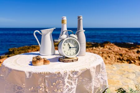Antique alarm clock next to white decoration bottles, on a round table overlooking the sea and...