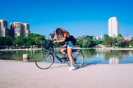 Valencia, Spain - July 22, 2019: A young tourist girl stops her rental bike in front of artificial lake in the old riverbed of the Turia river, converted into a garden in Valencia.のeditorial素材