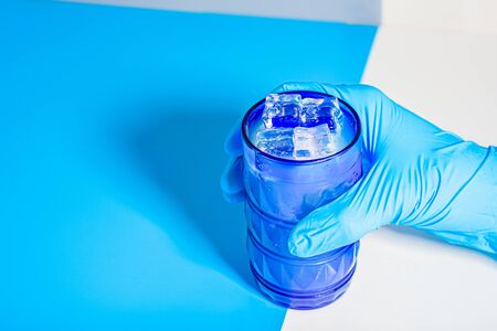 Hand of person with latex gloves holds a glass to drink it alone isolated in blue tones studio.