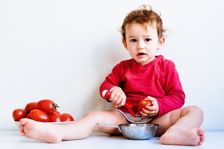 Baby looks at camera soiled with natural tomato juice while chewing vegetables, isolated on white.の写真素材