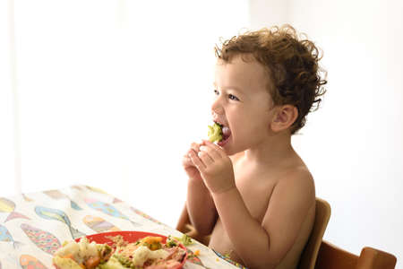 A young girl sitting in her high chair grabs a piece of vegetables and eats it by herself.の写真素材